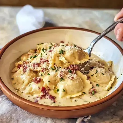 Eine Schüssel mit cremigen Ravioli, garniert mit Speckstückchen und Petersilie, mit einer Gabel, die etwas herausnimmt.