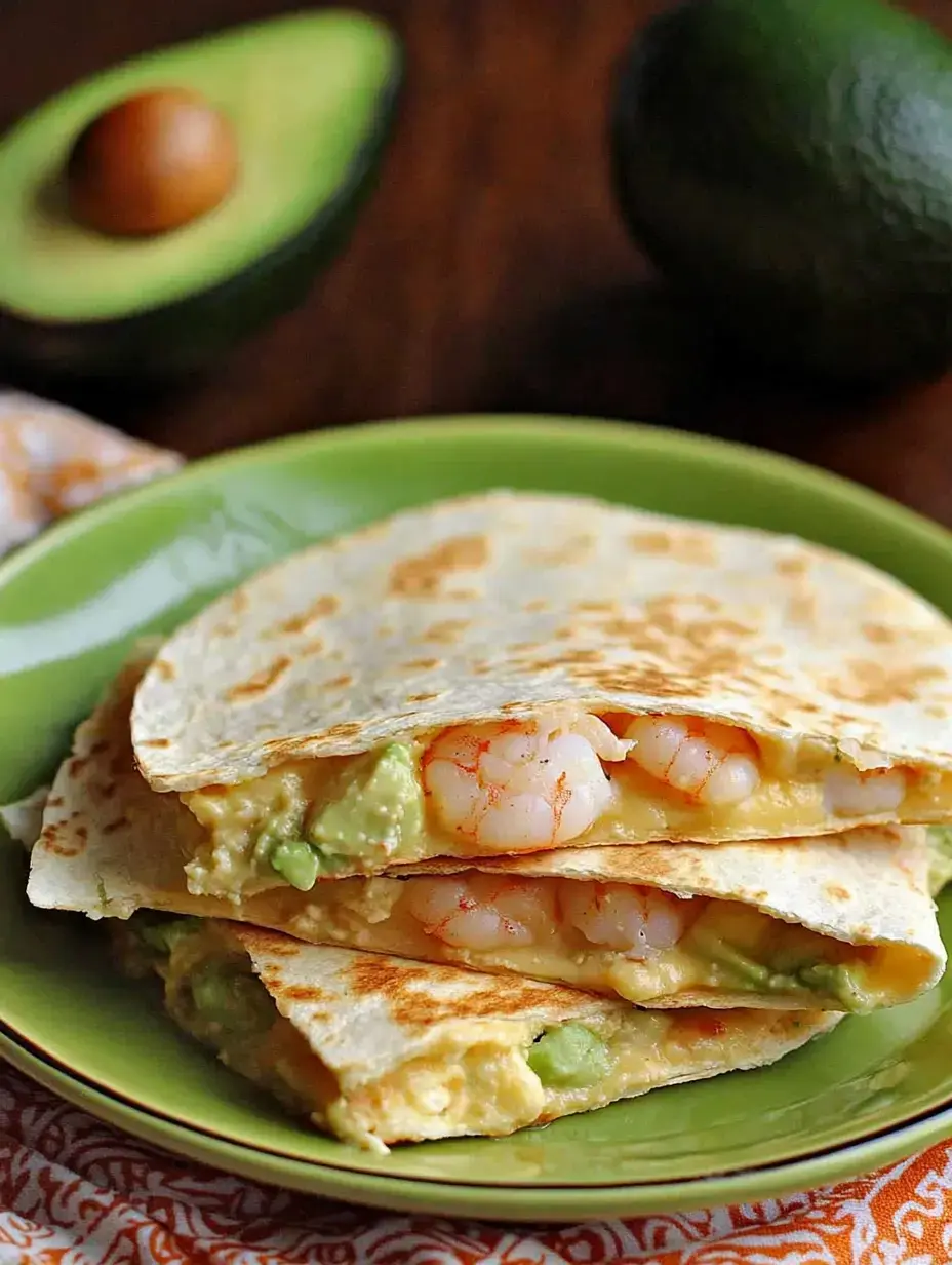 Eine Nahaufnahme eines grünen Tellers mit gestapelten Quesadillas gefüllt mit Garnelen und Avocado, begleitet von einer halbierten Avocado im Hintergrund.