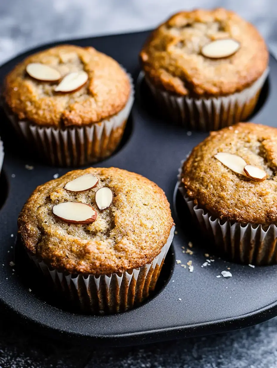 Vier frisch gebackene Mandelmuffins liegen in einer schwarzen Muffinform mit gehackten Mandeln obendrauf.