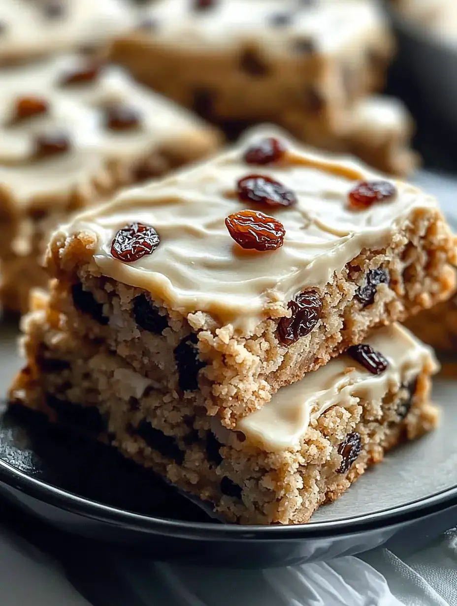 Eine Nahaufnahme von zwei gestapelten Stücken glasiertem Kuchen mit Rosinen obendrauf auf einem schwarzen Teller.