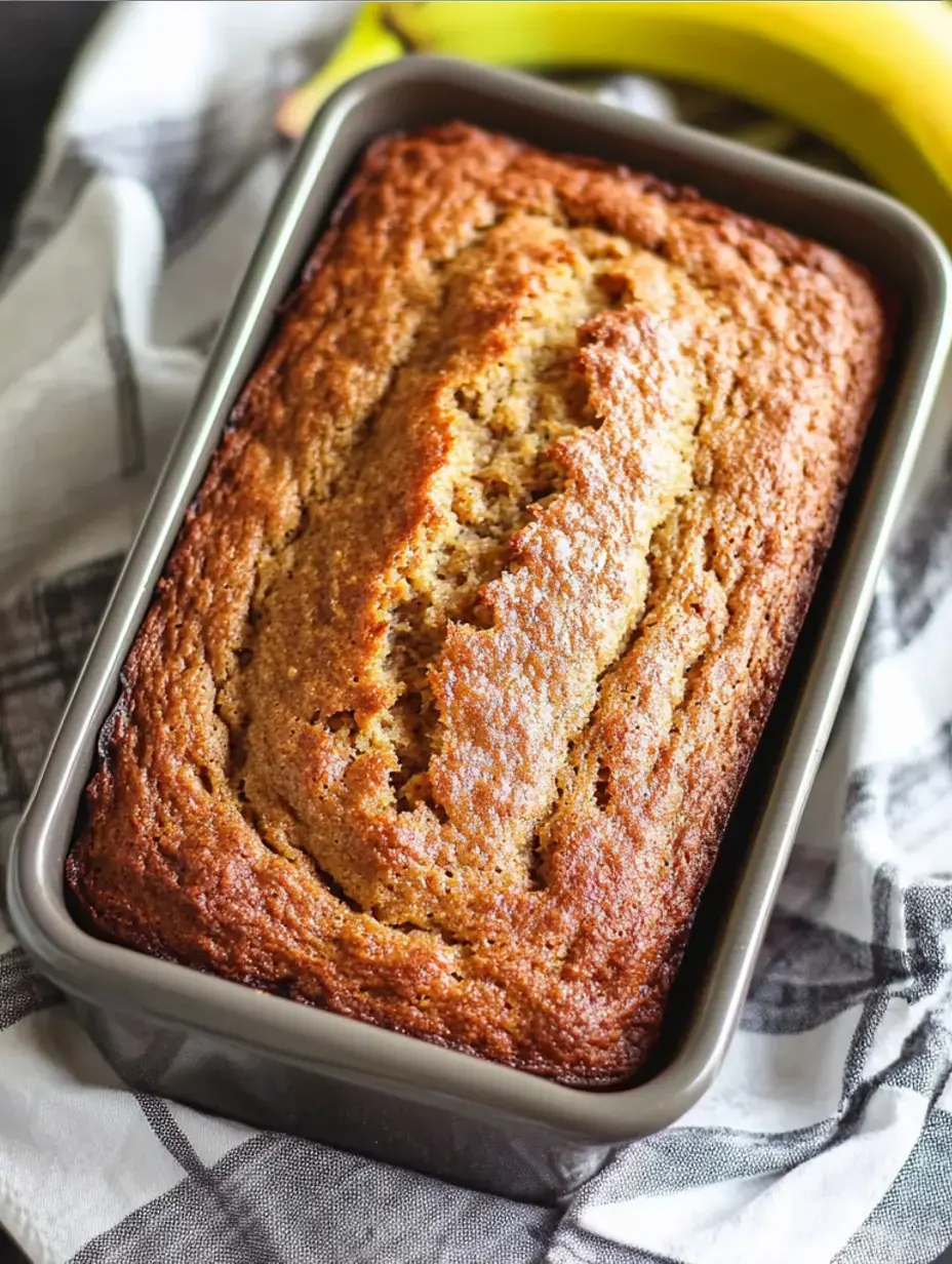 Ein frisch gebackener Laib Bananenbrot liegt in einer grauen Backform, umgeben von reifen Bananen und einem gemusterten Tuch.